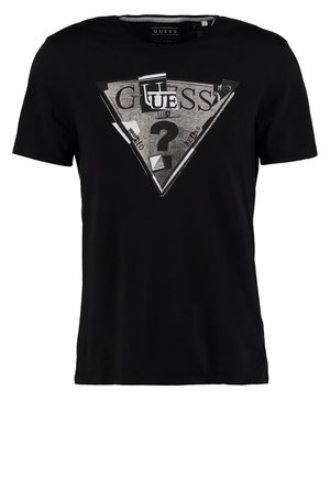 Mężczyzna w czarnej koszulce Guess, jasnoniebieskich obcisłych dżinsach, czarnych butach sportowych Adidas oraz z czarną torbą na ramię przewieszoną przez jedno ramię.