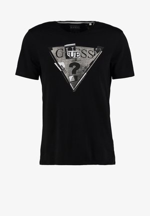 Man draagt een zwart Guess T-shirt, lichtblauwe strakke jeans, zwarte Adidas sneakers en heeft een zwarte crossbodytas over één schouder.