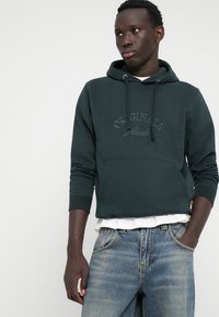 Haut à capuche vert foncé avec poche kangourou et texte « ORIGINALS » en relief. Porté sur une chemise blanche et assorti à un jean bleu délavé.