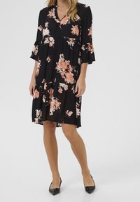 Femme portant une robe florale noire arrivant au genou avec des fleurs pêches et blanches et des escarpins à talon aiguille noirs.
