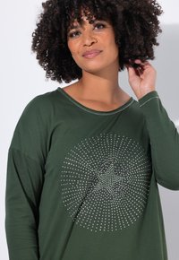 T-shirt vert à manches longues en tissu doux, arborant un motif circulaire de pierres à rhin brillantes au centre et un décolleté large.
