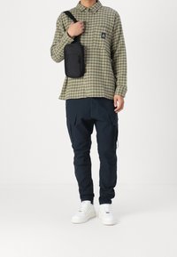 Grön- och brunrutigt långärmad skjorta, marinblå cargobyxor, vita sneakers och en svart axelväska med dragkedjeficka.