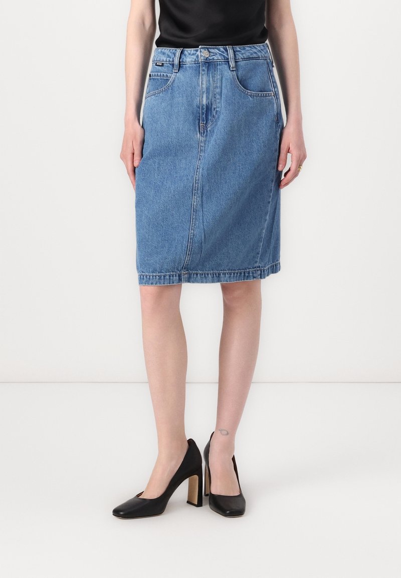 Falda lápiz de mezclilla en azul claro, hasta la rodilla, con una costura frontal, presillas para cinturón y cinco bolsillos. Combinada con zapatos de tacón alto negros.