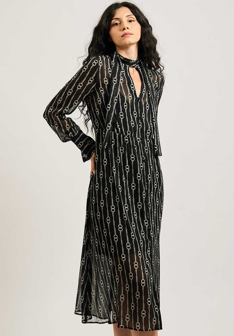 Abito nero trasparente con un motivo a catena verticale in oro. Maniche lunghe, dettaglio del collo a chiave e silhouette a forma di A.