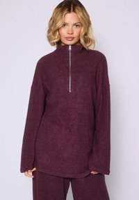 Sudadera de felpa burdeos con cremallera, que presenta un cuello alto, un ajuste relajado y mangas largas con dobladillo curvado. Textura suave con un detalle mínimo.