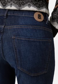 Tmavě modré džíny z denimu s hladkou texturou, standardními kapsami a hnědou koženou nášivkou se písmenem "B" na zadním pásku.