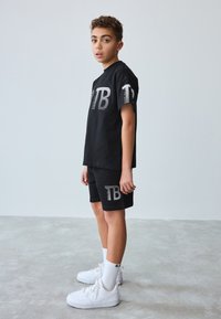 Sort bomulds-T-shirt med hvidstribet "TB"-logo, kombineret med sorte shorts med matchende logo, båret med hvide sneakers og sokker.