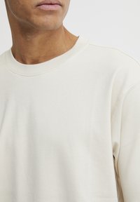 Lys beige genser med rund hals, glatt tekstur, synlige sømdetaljer og en avslappet passform, med fokus på komfort og enkelhet.