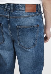Mørkeblå denimjeans med en brun skinnlapp i midjen, to baklommer med synlig søm og en rett bendesign.