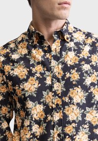 Blumiges Hemd mit Knöpfen, das einen navyfarbenen Hintergrund mit gelben und weißen Rosen, grünen Blättern und einem strukturierten Kordstoff zeigt. Schwarze Knöpfe sind enthalten.