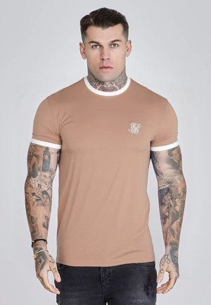 Man with tattoos on neck and arms wearing a fitted tan T-shirt with white trim, standing against a plain light background.
