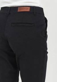 Close-up of a person wearing black pants with a back welt pocket and a brown leather "oxmo" brand patch on the waistband.