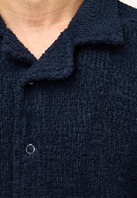 Camisa de color azul marino con textura, cuello con botones y un pequeño botón gris. La tela tiene una apariencia suave y tejida con una textura en bucles.