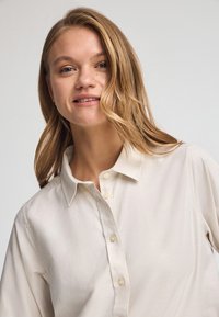 Jasno beżowa koszula zapinana na guziki wykonana z miękkiej tkaniny. Posiada klasyczny kołnierzyk oraz widoczne zapięcie na guziki. Luźny krój bez wzorów.