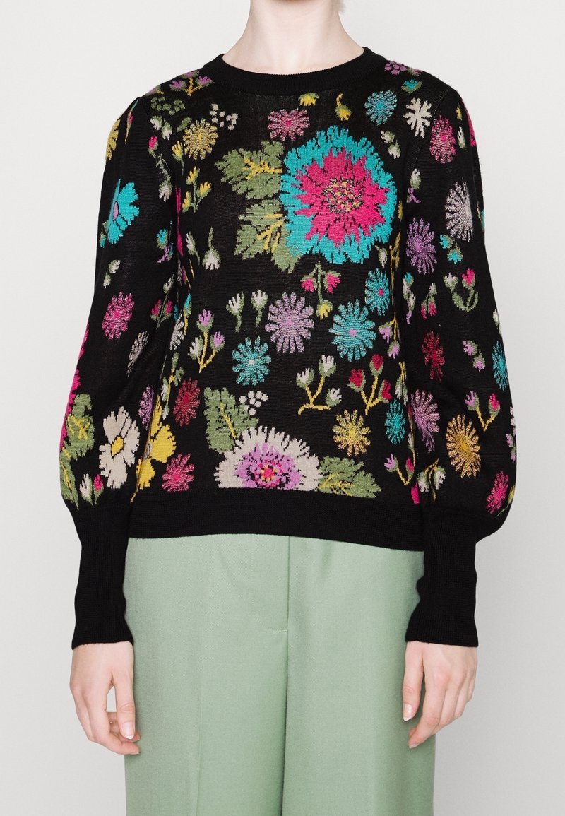 Pull noir à manches longues avec de grands motifs floraux colorés, associé à un pantalon taille haute vert clair porté par une personne.