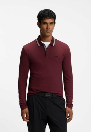 Hombre con cabello corto y rizado, vestido con una camisa polo de manga larga color burdeos, pantalones negros y cinturón negro, mirando hacia adelante.