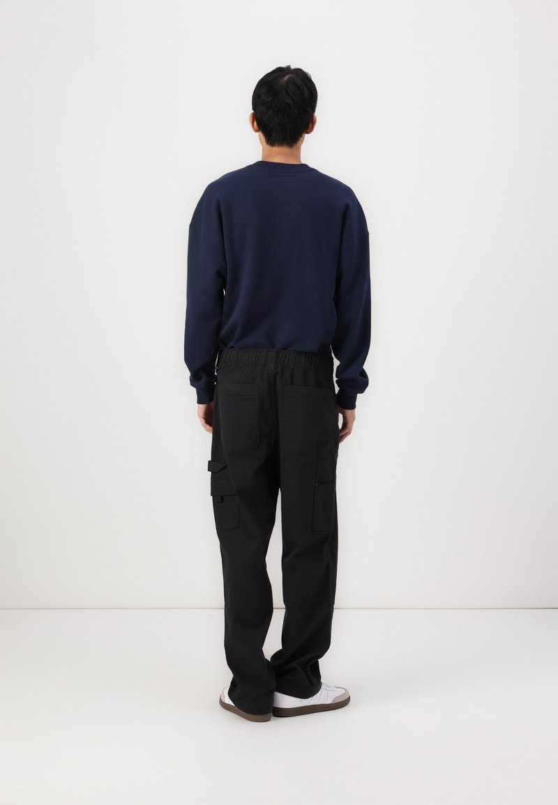 Felpa navy a maniche lunghe, abbinata a pantaloni cargo neri con tasche laterali. Il modello è di spalle contro uno sfondo neutro.