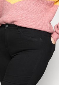 Personne portant un jean noir cintré et un pull en laine rose avec un détail jaune, une main dans la poche.