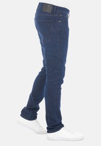 Vaqueros de mezclilla azul oscuro con un corte ajustado, diseño de cinco bolsillos y un parche de cuero en la cinturilla trasera, combinados con zapatillas blancas.