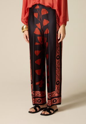 Donna che indossa pantaloni neri a gamba larga con motivi floreali rossi e bordi, camicetta rossa, braccialetti dorati e sandali neri a ciabatta, in piedi su un pavimento chiaro.