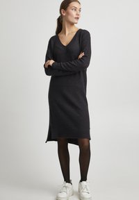 Svart stickad klänning med V-ringning, långa ärmar och sidofåror. Bärs med svarta, ogenomskinliga strumpbyxor och vita sneakers. Mjuk textur.