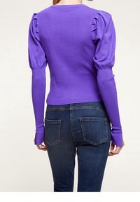 Frau in einem eng geschnittenen lila gerippten Pullover mit schwellenden langen Ärmeln und dunkelblauen Jeans, von hinten vor einem einfarbigen Hintergrund gezeigt.
