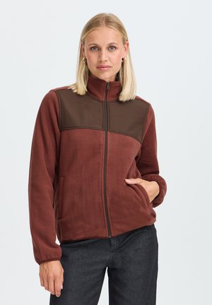 Blonde Frau mit Creolen und einer braunen Fleecejacke mit Reißverschluss sowie schwarzen Hosen, steht mit einer Hand in der Jackentasche.