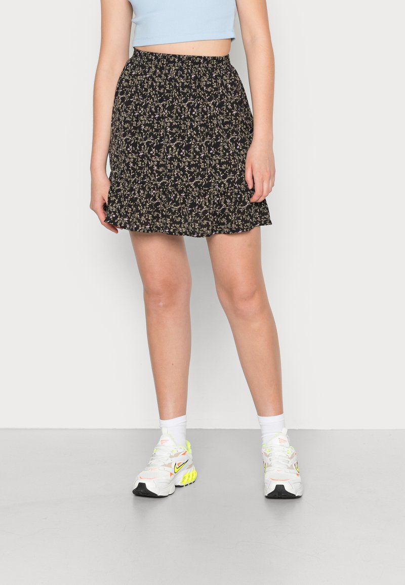 Saia preta floral com cintura franzida, apresentando pequenas flores roxas e amarelas. Usada com meias brancas e ténis.