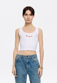Witte geribbelde crop top met rode geborduurde chilipeper en "Bimbay Lola" tekst. Het model combineert het met hooggesneden blauwe jeans.