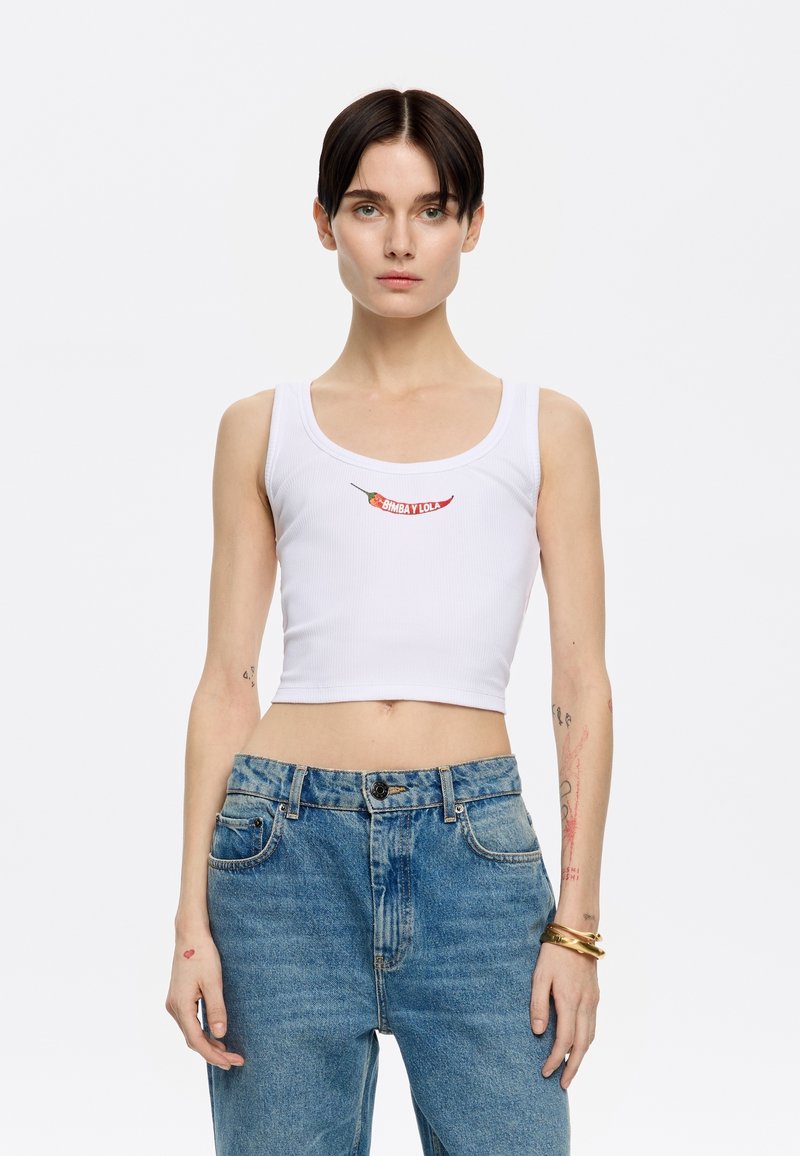 Witte geribbelde crop top met rode geborduurde chilipeper en "Bimbay Lola" tekst. Het model combineert het met hooggesneden blauwe jeans.