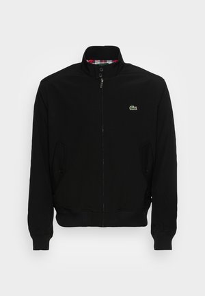 Uomo che indossa una giacca nera con zip e alto colletto, con il logo verde del coccodrillo sul petto, con le mani nelle tasche.