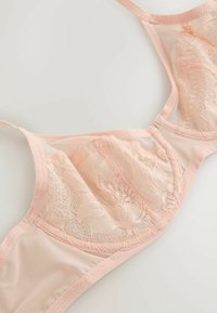 Lyserød blonder-bh med transparente mesh-stykker og bøjle, lagt fladt på en hvid overflade.