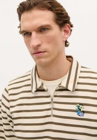 Polo shirt cu fermoar, cu dungi crem și maronii, decorat cu un design brodat ce include flori și un pește, guler rotund și textură moale.