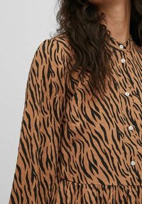 Kobieta w brązowej i czarnej sukience w paski tygrysie z długim rękawem, z białymi guzikami i długimi falującymi ciemnymi włosami widocznymi na ramieniu.