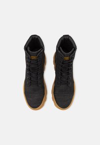 Baskets montantes noires en toile avec une finition texturée et une semelle en gomme. Présentent des lacets noirs et un petit logo jaune sur le talon.