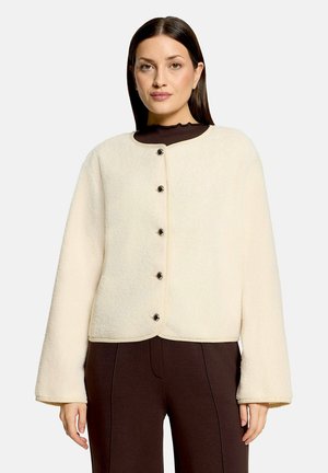 Frau mit glatten dunklen Haaren, die eine cremefarbene Fleecejacke mit Knöpfen und dunkelbraune Hosen trägt, steht vor einem schlichten Hintergrund.