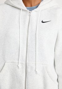 Nærbilde av en person som har på seg en lys grå hettegenser med glidelås, hette, snorer, frontlommer, og en liten svart Nike-logo på brystet.