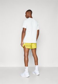 Camiseta blanca de manga corta con detalles desgastados, combinada con pantalones cortos amarillos brillantes con un patrón floral. Zapatillas deportivas y calcetines blancos.