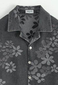 Chemise en denim noire à manches courtes, décorée d'un motif floral dans des nuances de gris, avec un col évasé et des boutons argentés sur le devant.