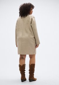Παλτό σε μπεζ από μείγμα μαλλιού με ίσια σιλουέτα, μακριά μανίκια και λεπτομέρεια ραφής στην πλάτη, συνδυασμένο με καφέ μπότες από σουέντ μέχρι το γόνατο.