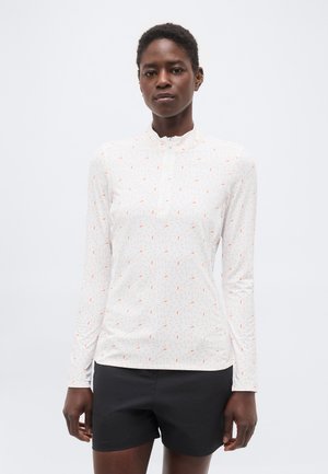 Žena v bielom dlhom rukáve s jemnými oranžovými a sivými bodkami a čiernymi šortkami stojaca pred jednoduchým svetlým pozadím.