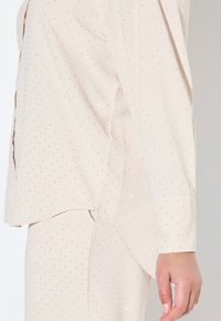 Top beige claro con lunar plateado brillante, mangas largas y un dobladillo ligeramente asimétrico. Fabricado con una tela suave y fluida.
