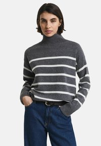 Pull gris rayé avec un col montant et des manches longues, présentant des rayures horizontales blanches et une texture tricot douce. Associé à un jean bleu.
