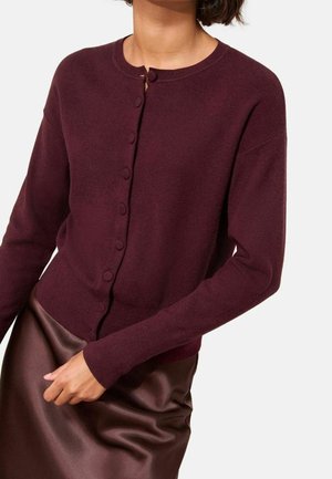 Kvinde iført en bordeaux knaplukket cardigan med lange ærmer kombineret med en matchende satin nederdel mod en ensfarvet baggrund.