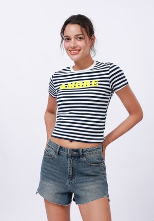 Top a strisce a maniche corte in blu navy e bianco con la scritta "AMORE" stampata in neon giallo; abbinato a pantaloni di jeans a vita alta con orlo sfrangiato.