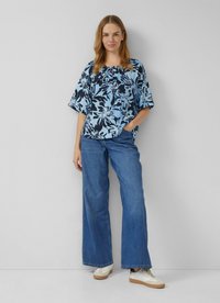 Frau steht in weit geschnittenen blauen Jeans, weißen Sneakers und einem lockeren blauen Blusenshirt mit Blumenmuster, die Hände in den Taschen, vor einer schlichten Wand.