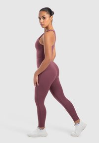 Bordeaux sportieve jumpsuit met dunne bandjes, nauwsluitend ontwerp en een gladde textuur, gecombineerd met witte sneakers met een logo op de enkel.