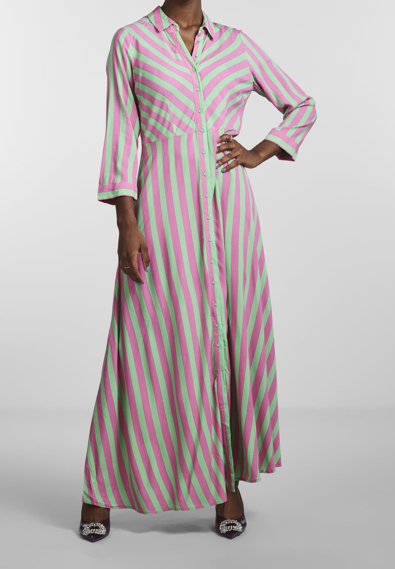 Femme portant une longue robe boutonnée avec des rayures diagonales roses et vertes et des manches trois-quarts, debout avec une main sur la hanche.