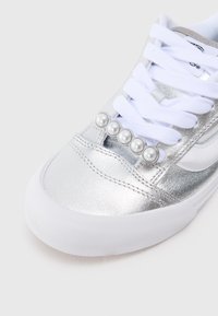 Pantofi sport metalici argintii cu sireturi albe, având cinci accente asemănătoare perlă pe partea superioară și o talpă din cauciuc alb pentru aderență.
