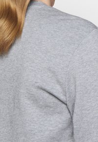 Sudadera gris con una superficie lisa y texturizada y un corte relajado. Presenta un cuello redondo y mangas sastreras, mostrando una parte del cabello.